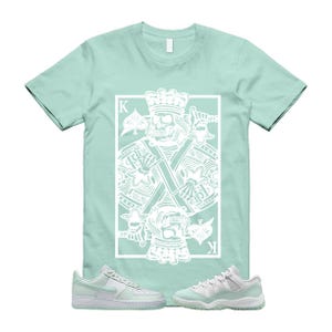 May include: A light green t-shirt with a white graphic of a skull king playing card design. The shirt is paired with light green and white sneakers. The sneakers are a low-top Air Force 1 and a pair of Jordan 11s.