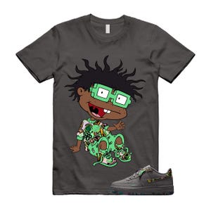 Pode incluir: Uma t-shirt cinzenta escura de manga curta com o gráfico de uma personagem de desenhos animados com cabelo preto espetado, óculos verdes e um conjunto verde estampado. Abaixo da t-shirt está um ténis cinzento escuro a combinar com um padrão no calcanhar.