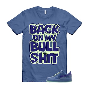 May include: Blue t-shirt with the text "BACK ON MY BULL SHIT" in a bold, outlined font. A blue sneaker is positioned at the bottom right. The shirt is a solid blue color.
