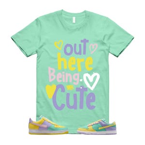 May include: A mint green t-shirt with the phrase "Out Here Being Cute" in colourful lettering and heart graphics. The image also features a pair of pastel-coloured trainers with yellow, purple, and light blue accents.