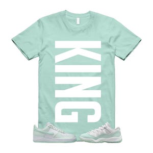 Puede incluir: Una camiseta verde menta con la palabra "KING" impresa verticalmente en blanco. La camiseta se combina con zapatillas deportivas verdes menta y blancas. Las zapatillas son de estilo bajo y alto.