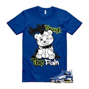 May include: Royal blue t-shirt featuring a distressed teddy bear graphic with the text "Smile Through The Pain" in black and yellow. A pair of blue, white, and yellow sneakers are visible in the image.