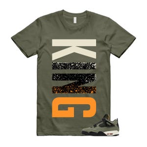 May include: Olive green t-shirt with the word "KING" in a bold, layered design. The "K" is cream, the "I" is black with white speckles, the "N" is black with orange speckles, and the "G" is orange. A matching olive green and black sneaker is at the bottom.