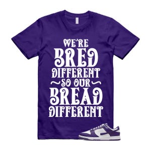 以下が含まれることがあります： 「WE'RE BRED DIFFERENT SO OUR BREAD DIFFERENT」の白文字がプリントされた紫色のTシャツ。白と紫のスニーカーが画像の右下にあります。