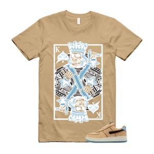 May include: Tan t-shirt featuring a playing card design with a skull king holding crossed swords in light blue and white. A tan and black sneaker with a blue sole is in the lower right corner.