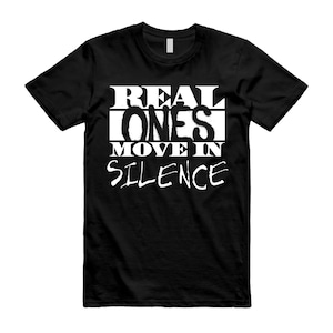 May include: Black t-shirt with the white text "REAL ONES MOVE IN SILENCE". The text is in a bold, stylized font. The shirt is a crew neck style.