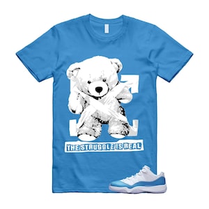 May include: A light blue t-shirt featuring a white teddy bear graphic with a white arrow design. The text "THE STRUGGLE IS REAL" is below the bear. A pair of white and light blue sneakers are at the bottom.