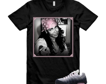 May include: A black short-sleeve t-shirt featuring a square graphic print of a person wearing a pink bandana, pointing forward. Below the graphic, a grey and black athletic sneaker with pink accents is displayed on the shirt's lower right side.