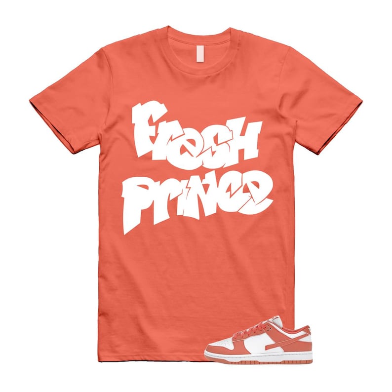 Puede incluir: Una camiseta de color coral con las palabras "Fresh Prince" en letras blancas grandes de estilo graffiti. Un par de zapatillas blancas y coral est&aacute;n en la parte inferior de la imagen. La camiseta tiene cuello redondo.
