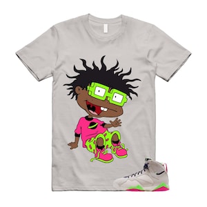 May include: A light gray short-sleeve t-shirt featuring a graphic of a cartoon character with spiky black hair, bright green glasses, a pink shirt with a black planet icon, and green shorts with pink spots, paired with a high-top sneaker.