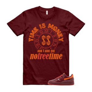 Könnte beinhalten: Burgunderfarbenes T-Shirt mit dem orangefarbenen Text "TIME IS MONEY" und einem Zifferblatt-Design mit Dollarzeichen. Darunter steht der Text "and I ain't got no freetime". Ein burgunderfarbener und orangefarbener Sneaker befindet sich unten.
