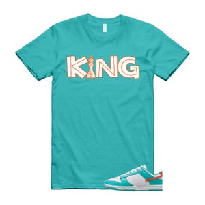 May include: A turquoise t-shirt with the word "KING" in white outlined in orange, featuring a chess piece. Below the shirt is a pair of white, turquoise, and orange trainers.