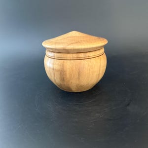 May include: A small, handcrafted wooden box with a lid. The box is light brown with a rounded body and a conical lid. The lid fits snugly on the box, and there are two thin, dark lines around the middle of the box. The box is sitting on a dark surface.