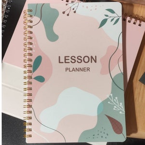 May include: A pink and green spiral bound notebook with the text "LESSON PLANNER" on the cover. The cover has a floral design with abstract shapes.