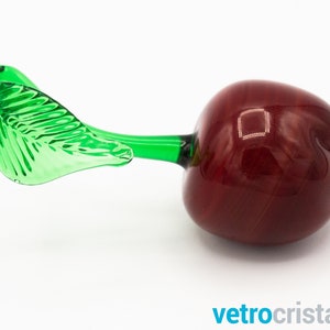 May include: A glass cherry with a green leaf stem. The cherry is a deep red color and the leaf is a bright green.