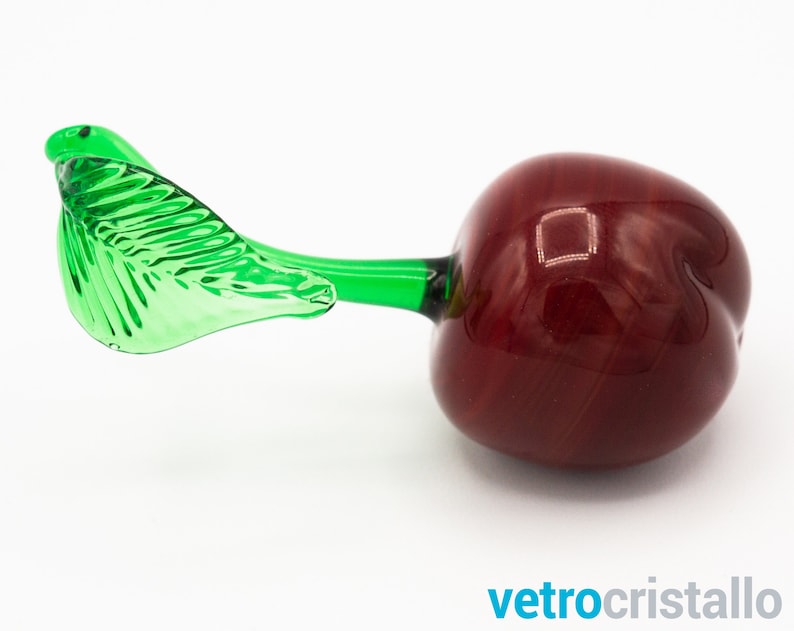 May include: A glass cherry with a green leaf stem. The cherry is a deep red color and the leaf is a bright green.