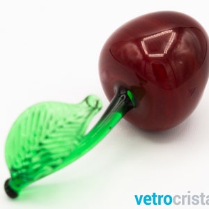 May include: A glass cherry figurine with a green leaf stem. The cherry is a deep red color and the leaf is a bright green.
