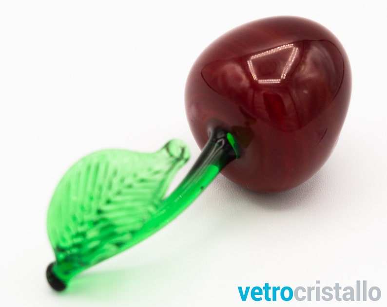 May include: A glass cherry figurine with a green leaf stem. The cherry is a deep red color and the leaf is a bright green.