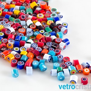 May include: A collection of colorful, cylindrical glass beads with intricate floral and geometric patterns. The beads are in various colors, including red, blue, orange, and white, scattered on a white surface. The image includes the text "vetrocristallo".