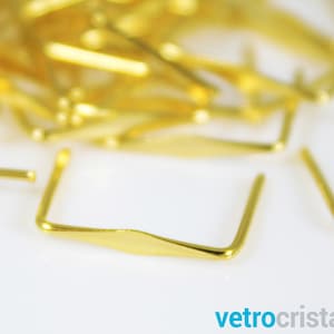 May include: A close-up of a gold metal U-shaped clasp with a flat, diamond-shaped center. The clasp is lying on a white surface.