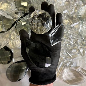 May include: A black gloved hand holds a clear, faceted crystal ball, a black teardrop-shaped crystal, and a clear, triangular crystal. The background is filled with various clear and faceted crystal shapes, suggesting a collection of decorative crystals.
