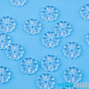 May include: Clear, flower-shaped glass beads with a central hole are scattered on a light blue surface. The beads have a delicate, translucent appearance, with petal-like edges. The overall effect is one of simple elegance and charm.
