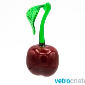May include: A glass cherry figurine with a green stem and leaf. The cherry is a deep red color and has a glossy finish.