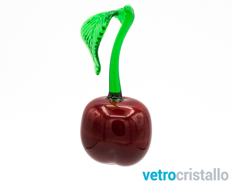 May include: A glass cherry figurine with a green stem and leaf. The cherry is a deep red color and has a glossy finish.