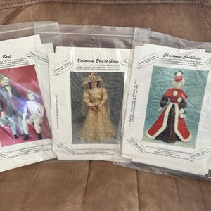 May include: Three sealed plastic bags containing sewing patterns. The patterns feature images of dolls in period clothing, including a Victorian bridal gown, a Christmas coatdress, and a man and boy in formal attire. The patterns are on white paper.