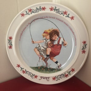 May include: A white ceramic plate featuring a vintage illustration of two children jumping rope. The plate has a floral border with the word "JUNE" and text that reads "A certified plate in the original and only Sarah Stilwell Weber Calendar Collection."