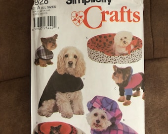 May include: Simplicity Crafts sewing pattern for dog clothes and beds. The pattern features illustrations of dogs wearing various outfits and resting in beds. The pattern includes the text "928" and "Crafts".