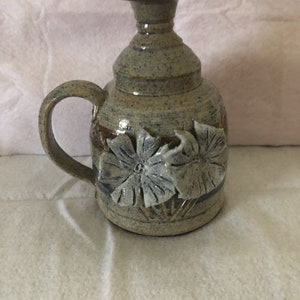 May include: A handcrafted ceramic pitcher with a handle and a flared top. The pitcher is a speckled gray-blue with a brown band and two floral designs. The flowers are a lighter blue-gray. The pitcher is a unique piece of pottery.