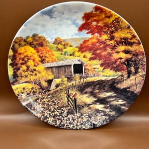 May include: A decorative plate featuring a scenic autumn landscape with a covered bridge, trees in vibrant shades of red, orange, and yellow, and a blue sky. The plate has a glossy finish and the artist's signature, "JERNER", is visible.