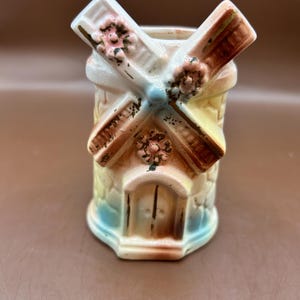 May include: A ceramic windmill figurine with a detailed design. The windmill features a white and brown rotating fan, a stone-like structure, and a blue and yellow color scheme. Small pink floral accents are present.
