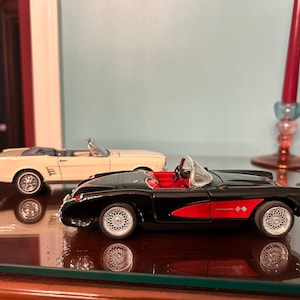 May include: Two model cars: a black convertible with red accents and a cream-colored convertible. The black car has silver wheels and a red interior. The cream car is in the background. Both cars are on a reflective surface.