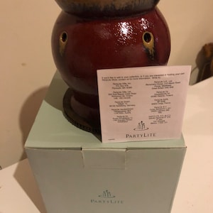 May include: A burgundy ceramic PartyLite oil warmer with a dark brown rim and two side openings. The warmer sits on a light blue box with the PartyLite logo. A white paper with text is placed on the box.