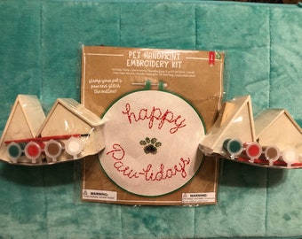 May include: A Pet Handprint Embroidery Kit with a stitched design that says "happy paw-lidays". The kit includes a canvas hoop, needle, thread, and paint. Two small wooden houses with paint pots are on either side.