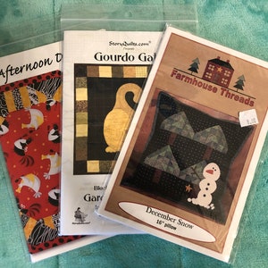 May include: Three packaged quilt patterns. One is titled "Afternoon D" with a red and black pattern. Another is "Gourdo Gal" with a goose design. The third is "December Snow" with a snowman and tree design, for a 16-inch pillow.