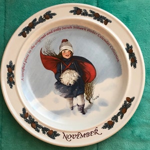 May include: A white ceramic plate from the Sarah Stilwell Weber Calendar Collection. The plate features a young person in a winter scene, wearing a red cape and holding a white bundle. The word "November" is at the bottom.