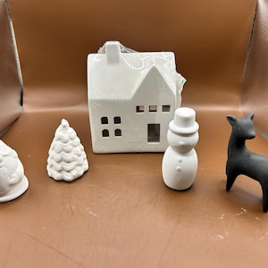 May include: A collection of ceramic holiday decorations, including a white house with cut-out windows, a snowman, a gnome, a Christmas tree, and a black deer. These items are unpainted and ready for customization.