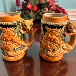 May include: Two ceramic beer steins with a detailed relief design. Each stein features a deer and a squirrel-like animal as the handle. The steins are primarily orange, brown, and green, with a glossy finish. The steins are approximately 4 inches tall.