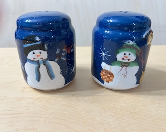 May include: A pair of cylindrical ceramic salt and pepper shakers with a glossy dark blue finish. Each shaker features a hand-painted snowman design with white snow, stars, and colorful winter accessories like scarves and hats.