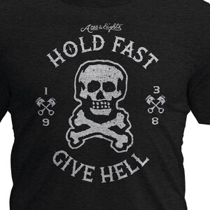 May include: Black crew neck t-shirt with a distressed white graphic. The graphic features a skull and crossbones with the words "HOLD FAST GIVE HELL" and the numbers "1938" and "1939". The shirt is made of a soft, heathered fabric.