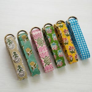 May include: Six colorful fabric straps with brass rings. The straps feature various patterns, including floral designs and a blue and white gingham check. The colors include tan, green, pink, yellow, and blue.