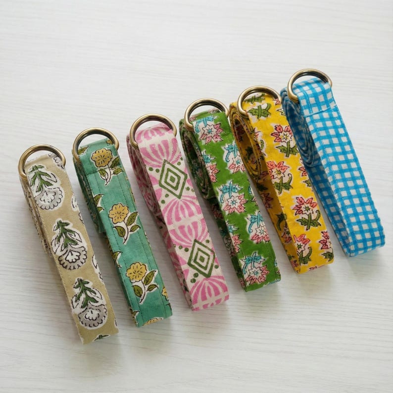 May include: Six colorful fabric straps with brass rings. The straps feature various patterns, including floral designs and a blue and white gingham check. The colors include tan, green, pink, yellow, and blue.