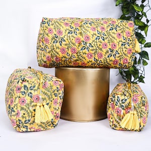 Può includere: Tre trousse per cosmetici e un cuscino cilindrico con stampa floreale gialla. Le borse e il cuscino presentano un motivo ripetuto di fiori rosa e foglie verdi su sfondo giallo. Ogni borsa ha un nappa gialla. Gli articoli sono esposti su un supporto dorato.