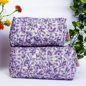 May include: Two stacked, rectangular cosmetic bags with a floral pattern in shades of purple and blue on a light pink background. Each bag has a matching tassel zipper pull. The bags are quilted.