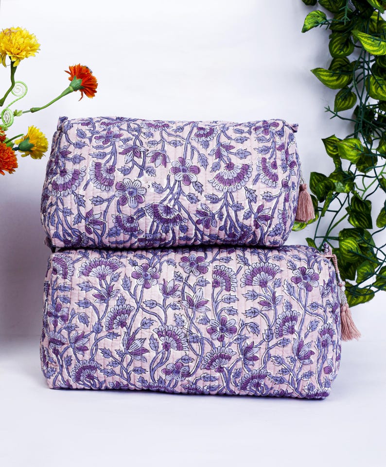 May include: Two stacked, rectangular cosmetic bags with a floral pattern in shades of purple and blue on a light pink background. Each bag has a matching tassel zipper pull. The bags are quilted.