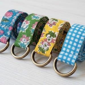 May include: Four fabric belts with brass D-rings. The belts feature floral and gingham patterns in blue, green, and yellow. The blue belt has a floral pattern, the green belt has a floral pattern, the yellow belt has a floral pattern, and the last belt is blue and white gingham.