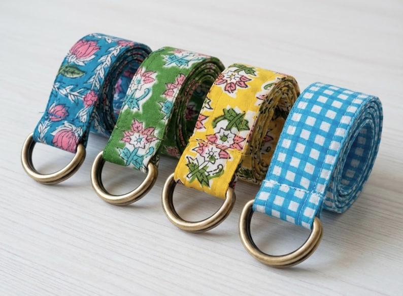 May include: Four fabric belts with brass D-rings. The belts feature floral and gingham patterns in blue, green, and yellow. The blue belt has a floral pattern, the green belt has a floral pattern, the yellow belt has a floral pattern, and the last belt is blue and white gingham.